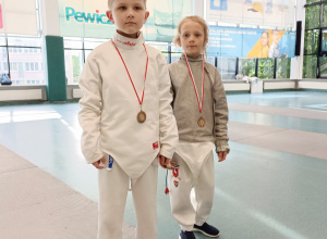 Dwoje uczniów stoi w sali sportowej w strojach szermierczych. Na szyi mają powieszone medale.