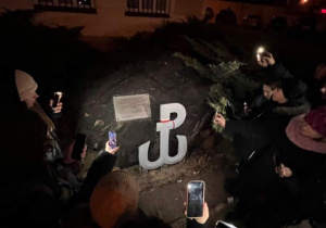 Młodzież stoi przy pomniku. Oświetla znak Polski Walczącej. To miejsce, w którym odbyła się akcja pod Arsenałem.