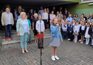 Uczennica śpiewa piosenkę na placu apelowym przed szkołą. W tle stoją panie dyrektor i rodzice uczniów.