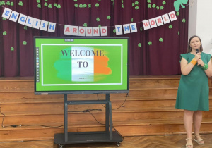 Nauczycielka uroczyście rozpoczyna Międzyszkolny Turniej Języka angielskiego. Po środku sali stoi monitor interaktywny. Za monitorem rozwieszona dekoracja z napisem Ireland.