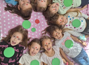 Na dywanie w kropki leży grupa dziewczynek. Każda z nich trzyma w dłoniach zieloną kropkę.