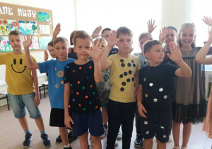 W Sali lekcyjnej grupa uczniów ubrana w koszulki w kropki. W dłoniach trzymają wykonane samodzielnie prace plastyczne.