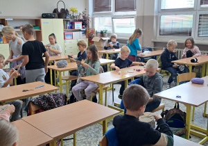 Sala lekcyjna. Uczniowie siedzą przy biurkach. Mają w ręku włóczkę, wykonują ozdoby. Troje uczniów chodzi między ławkami, udziela instruktażu.