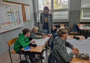 Uczniowie pracują przy stolikach w parach. Zapisują na plakatach pomysły na ochronę środowiska. Obok uczniów stoi starszy kolega, który pomaga w wykonaniu zadania.