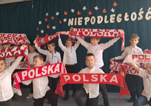 Grupa chłopców ubranych na galowo i trzymających w dłoniach czerwono – białe szaliki stoi na tle napisu: Niepodległość.