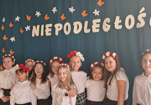 Grupa dziewczynek w biało – czerwonych opaskach na włosach stoi na tle napisu :Niepodległość.