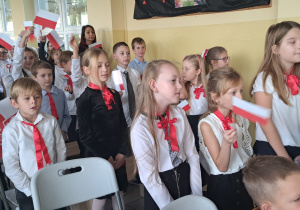 Duża grupa dzieci stoi na korytarzu szkolnym. Ubrani są na galowo, w rękach trzymają biało – czerwone flagi.