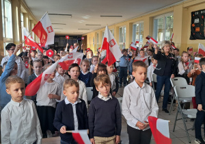 Grupa dzieci stoi na korytarzu szkolnym. Ubrani są na galowo, w rękach trzymają biało – czerwone flagi.