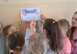 Grupa dziewczynek stoi przy ścianie. Na ścianie przyklejona jest kartka z napisem: ”Weź ze sobą dobre słowo”.