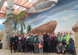 Uczniowie klas 6a i 6c pozują do zdjęcia w sali Afrykarium. W tle stoją palmy a nad głowami dzieci wisi delfin.