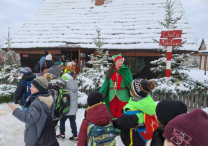 Grupa dzieci wchodzi do zaśnieżonej chaty Mikołaja. Towarzyszy jej kobieta przebrana w strój elfa.