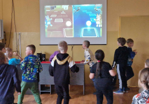 Dzieci uczestniczą w zajęciach ruchowych przed ekranem rzutnika. Na ekranie bohaterowie bajki "Bluey" oraz napis "Jump!" (Skacz!)