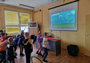 Dzieci uczestniczą w zajęciach ruchowych przed ekranem rzutnika. Na ekranie bańka mydlana. Dzieci wskazują bańkę.