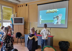 Dzieci uczestniczą w zajęciach na spostrzegawczość przed ekranem rzutnika. Na ekranie skomplikowany obrazek. Dzieci pokazują palcami odnaleziony szczegół.