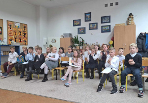 W sali lekcyjnej siedzi duża grupa dzieci odświętnie ubranych. W tle widać wyposażenie klasy.