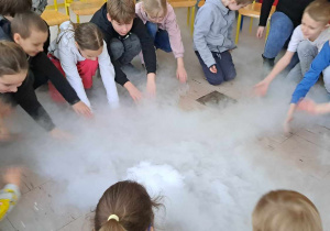 Duża grupa uczniów siedzi w kręgu. Pośrodku kręgu widać kłęby dymu powstałe dzięki suchemu lodowi.