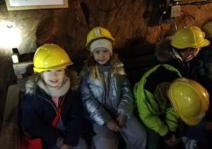 Grupa uczniów siedzi na ławce w podziemnym korytarzu kopalni. Na głowach mają żółte kaski.