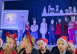 Na scenie widać grupę śpiewających uczniów. Na pierwszym planie grupa dzieci siedzi na podłodze. W tle dekoracje świąteczne.