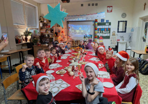 Duża grupa dzieci siedzi wokół świątecznego stołu. Na głowach mają czapki Mikołaja.