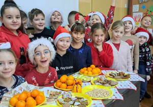 Grupa dzieci stoi wokół stołu. Na stole są talerze z mandarynkami oraz piernikami świątecznym