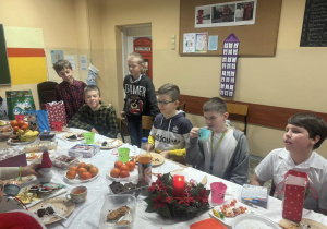 Uczniowie siedzą przy stole. Na stole jest stroik świąteczny, owoce i pierniki.