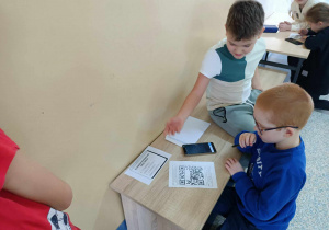Dwóch chłopców siedzi na ławce na szkolnym korytarzu. Przed sobą mają telefon i kartki z kodem QR. W tle widać pozostałych uczniów.