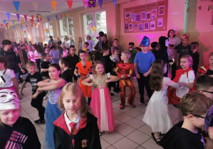 Duża grupa dzieci przebranych w stroje karnawałowe bawi się na holu. Uczniowie tańczą w rytm muzyki.