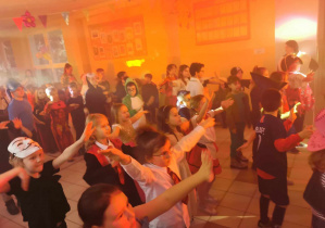 Duża grupa dzieci przebranych w stroje karnawałowe bawi się na holu. Uczniowie tańczą w rytm muzyki.