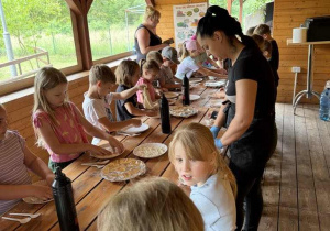 Przy długim stole stoi duża grupa dzieci. Na stole stoją talerze z ciastem na pizzę. Na końcu stołu kobieta tłumaczy dzieciom, jak wyrabiać ciasto na pizzę.