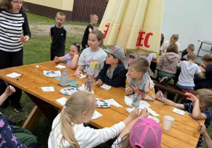 Na dworze, przy drewnianym stole siedzi grupa dzieci. Wszystkie mają w dłoniach pędzle i malują kamienie. Na stole widać rozłożone palety i słoiki z wodą.