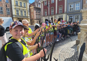 Duża grupa dzieci stoi wokół pomnika. W tle widać zabytkowe kamienice.
