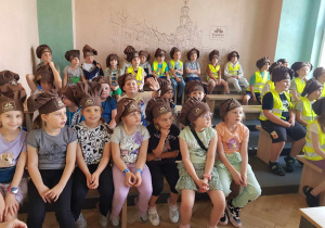 Duża grupa dzieci ubranych w odblaskowe kamizelki i czapki kucharskie siedzi w pomieszczeniu. Na ścianie widać narysowaną zabudowę miejską.