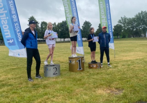 Uczennice 4 klas na podium z otrzymanymi medalami i nagrodami. Po obu stronach podium panowie, którzy wręczali medale. W tle chorągiewki z logiem MOSIR i stadion.