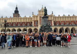 Grupa uczniów stoi pod pomnikiem. W tle ulica Krakowa i Sukiennice.
