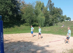 Trzech uczniów na piaszczystym boisku rozgrywa mecz piłki siatkowej. W tle widać zielone drzewa i chłopca, który siedzi na trawie i kibicuje grającej drużynie.