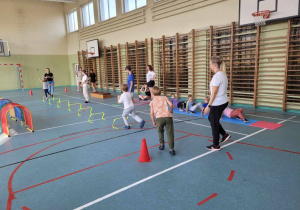 Dzieci kolejno wykonują polecenia nauczycielki. Biegną przeskakując przez przeszkody. Początek wyznaczają czerwone pachołki ustawione na drewnianej, zielonej podłodze.