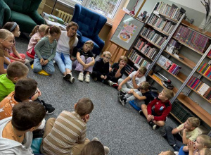 Uczniowie siedzą na szarym dywanie, słuchają bibliotekarki. Po prawej stronie stoi regał z książkami. W tle widać okno, pod którym stoją 2 fotele (granatowy i zielony) i stolik z lampą.