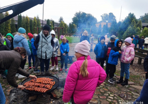 Uczniowie w kolorowych kurtkach i czapkach stoją wokół przygotowanego ogniska. Na okrągłym ruszcie nad ogniskiem ułożone są kiełbaski. Pani w brązowej kurtce piecze kiełbaski.