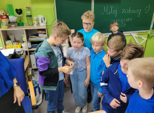 Uczniowie ubrani w stroje w kolorze niebieskim rozwiązują zagadkę. Chłopiec trzyma woreczek z zadaniem. W tle widać tablice, na której napisane jest: Miłej zabawy! Otrzęsiny 2024.