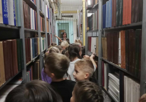 Grupa uczniów chodzi między pólkami w bibliotece. Pólki wypełnione są kolorowymi książkami.