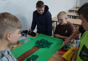 Uczniowie znajdują się w sali lekcyjnej. N ławce leży zielona karta papieru. Uczniowie wykonują plakat. Na jego środku widać drzewo wykonane z bibuły.