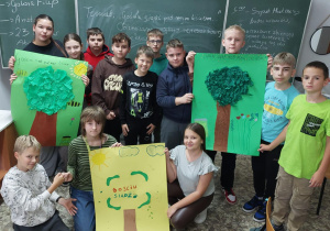 Uczniowie prezentują wykonane na lekcji plakaty. Widać trzy plakaty (dwa na zielonym tle, jeden na żółtym tle). Na środku każdej pracy znajduje się duże drzewo i hasło reklamowe.