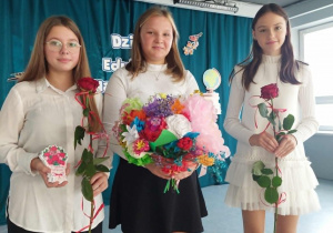Trzy uczennice trzymają prezenty dla nauczycieli: bukiet z kwiatków z bibuły, kartki z życzeniami i czerwone róże.