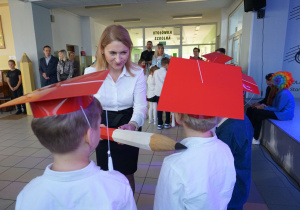 Uczniowie w białych koszulach i czerwonych biretach stoją przed Panią Dyrektor, która dokonuje pasowania dużą biało-czerwoną kredką.