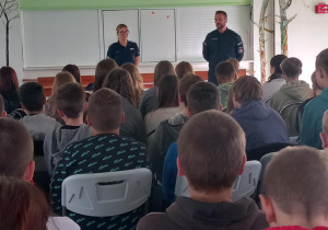 Policjant i policjantka prowadzą spotkanie z uczniami. Uczniowie siedzą na krzesłach. W tle widać białą tablicę.