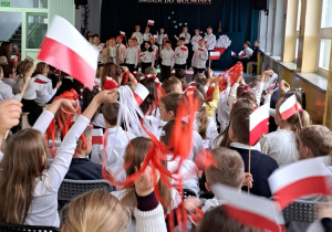 Uczniowie machają biało-czerwonymi flagami. W tle widać scenę i uczniów tam występujących.