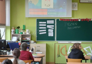 Przedstawiciele klas siedzą w sali lekcyjnej w ławkach i rozwiązują zadania wyświetlone na białej tablicy przed nimi.