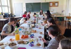 Uczniowie siedzą po obu stronach nakrytego stołu. Na stole znajdują się tradycyjne potrawy wigilijne i słodycze.