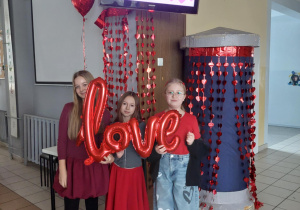 Uczennice trzymają w ręku balon z napisem love. Za nimi widać girlandę z serc, nad nimi wyświetlane są życzenia walentynkowe.