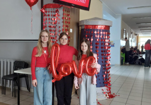 Uczennice trzymają w ręku balon z napisem love. Za nimi widać girlandę z serc, nad nimi wyświetlane są życzenia walentynkowe.